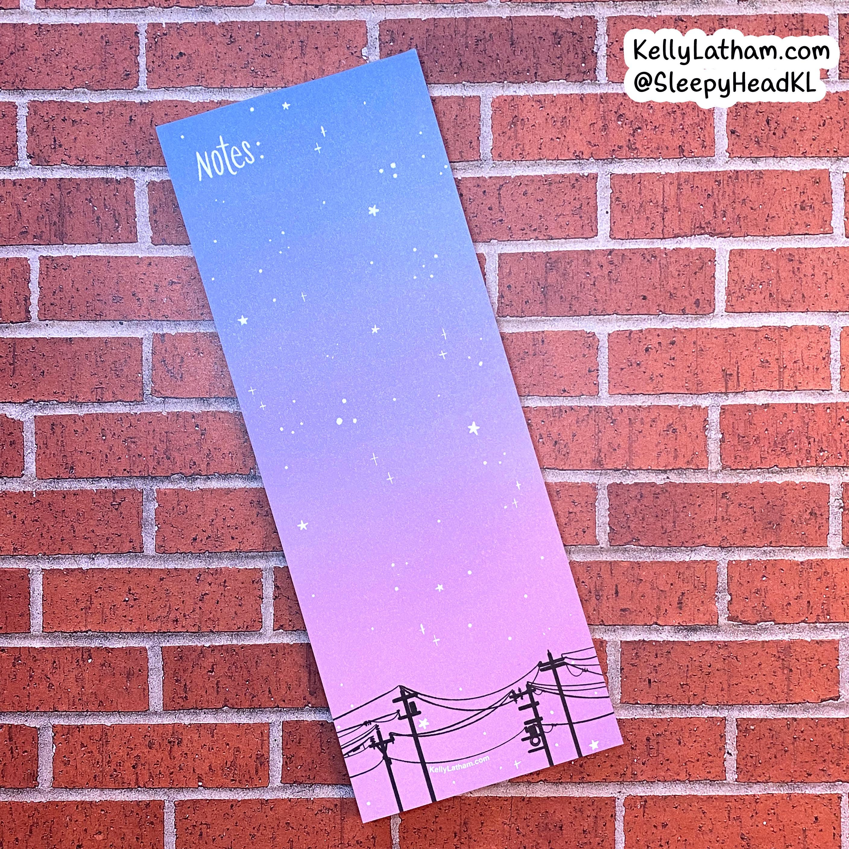 A vertical notepad with a blue-to-pink gradient sky and starry night design, featuring black utility pole illustrations at the bottom and the word 'Notes:' in the top left. The notepad is photographed against a red brick wall, with a brand watermark in the top right corner.