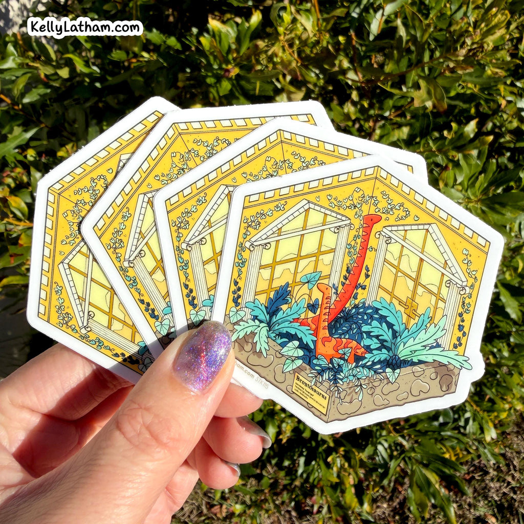 A hand with glittery purple nail polish holds a fan of five die-cut stickers featuring an illustration of a dinosaur inside a greenhouse, set against a background of green outdoor foliage. The watermark 'KellyLatham.