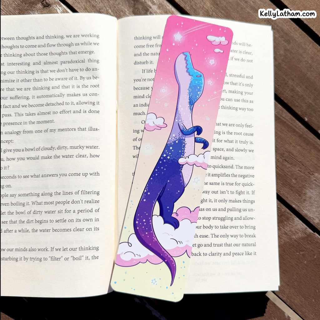 An open book with printed text spread open, decorated with a large pink and blue watercolor-style illustration of a mermaid with a pink tail, resting on a wooden surface.