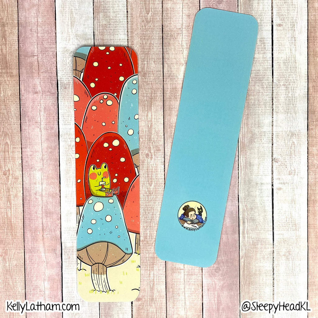 Two rectangular bookmarks or cardstock pieces displayed on a whitewashed wood plank background. One piece is plain light blue with a small circular cartoon sticker near the bottom, and the other shows a whimsical mushroom illustration in red, blue, yellow, and beige tones.