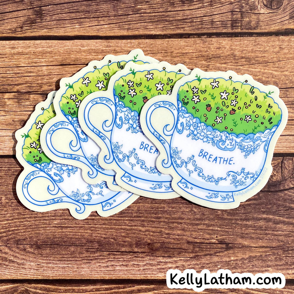Four die-cut stickers shaped like blue-and-white teacups with green grass and small flowers, arranged on a brown wood surface. One sticker shows the word 'BREATHE.