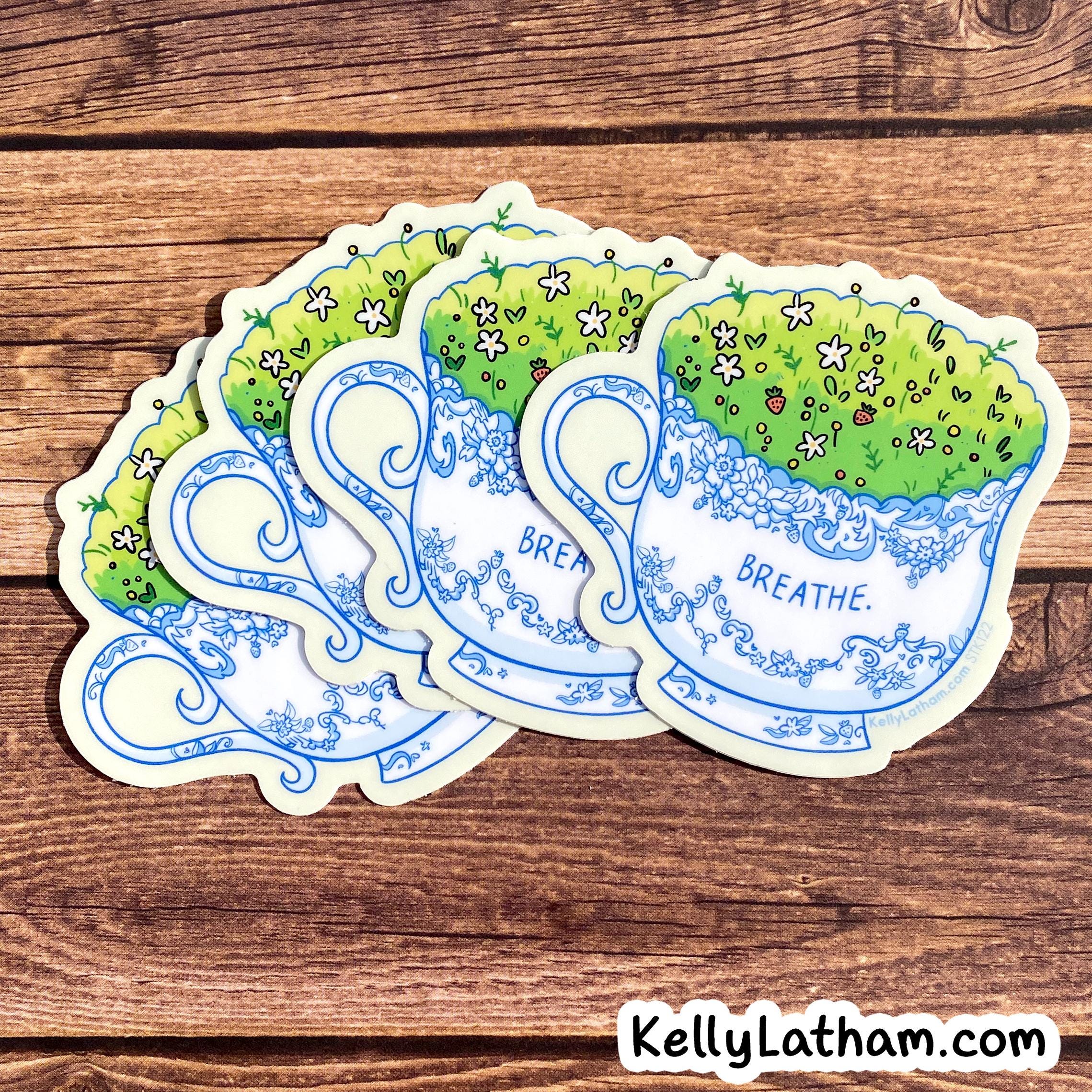 Four die-cut stickers shaped like blue-and-white teacups with green grass and small flowers, arranged on a brown wood surface. One sticker shows the word 'BREATHE.