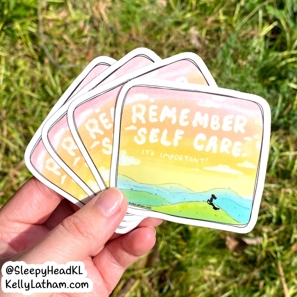 A hand holds a fanned-out set of square stickers outdoors on grass. The stickers have rounded corners and read “REMEMBER SELF CARE.