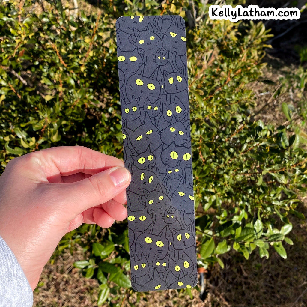 A hand holds a long, narrow dark gray card or coaster featuring a repeating pattern of black cats with glowing yellow eyes, set against a background of green garden foliage.