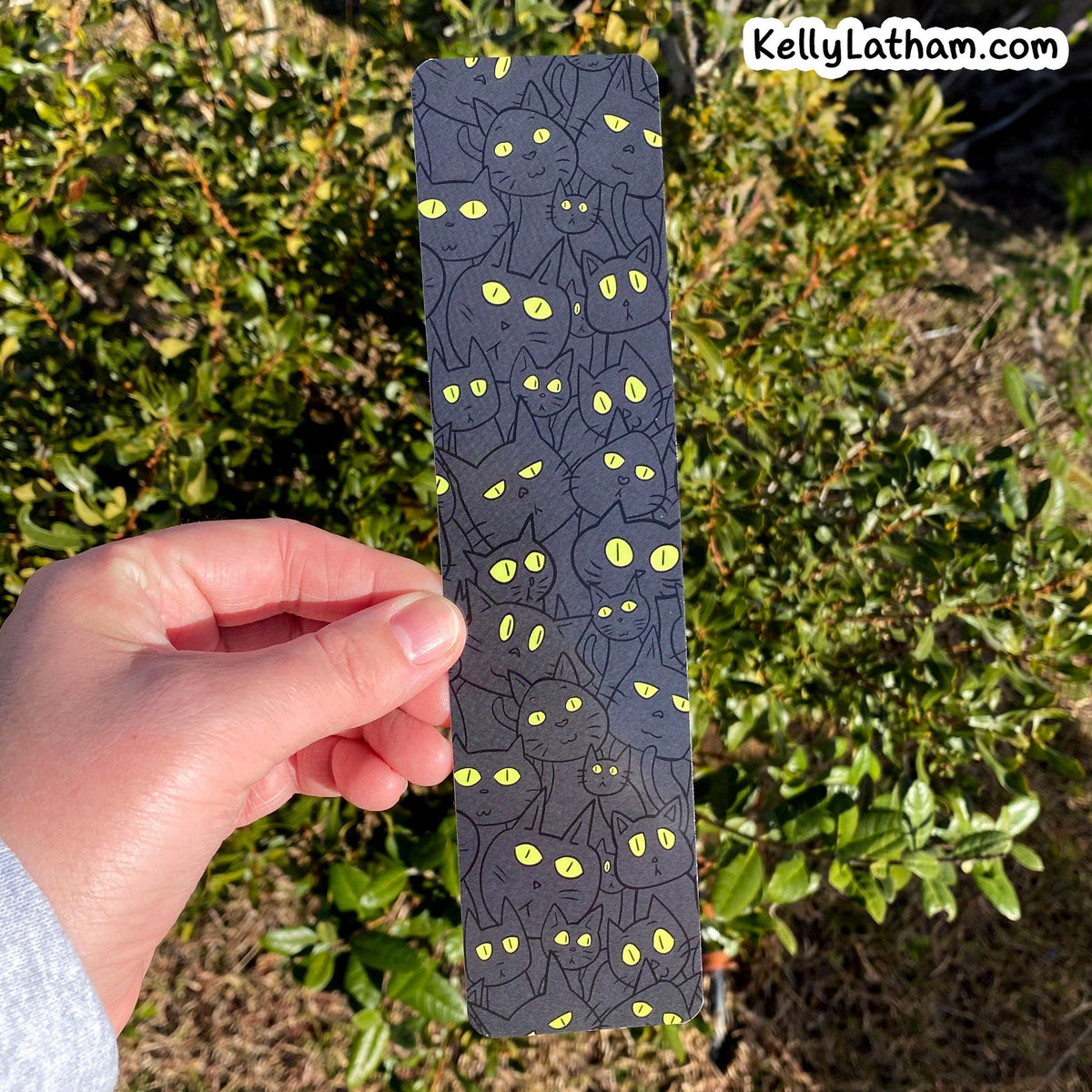 A hand holds a long, narrow dark gray card or coaster featuring a repeating pattern of black cats with glowing yellow eyes, set against a background of green garden foliage.