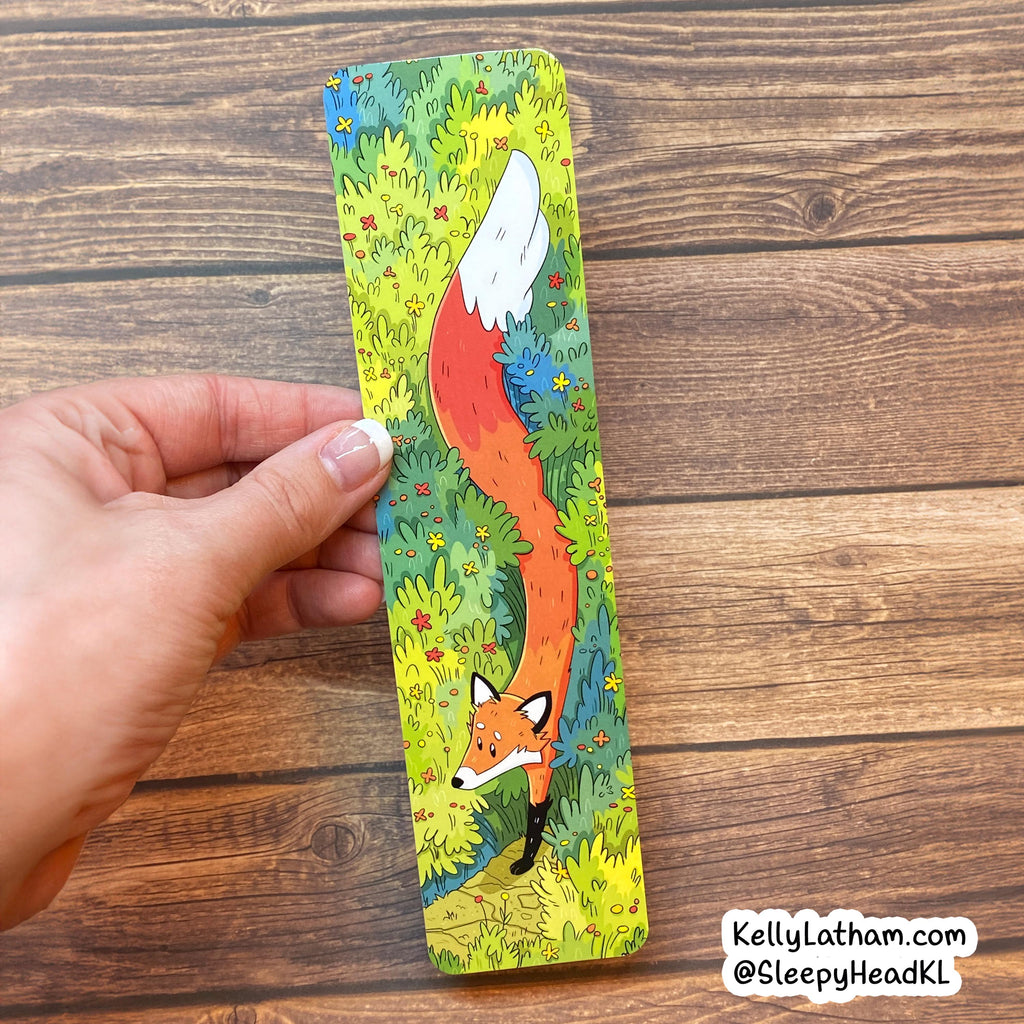 A hand holds a long rectangular printed card or bookmark featuring a whimsical fox illustration in a grassy field. The card has a colorful printed design with green, yellow, blue, orange, and white tones, photographed over a brown wood surface.