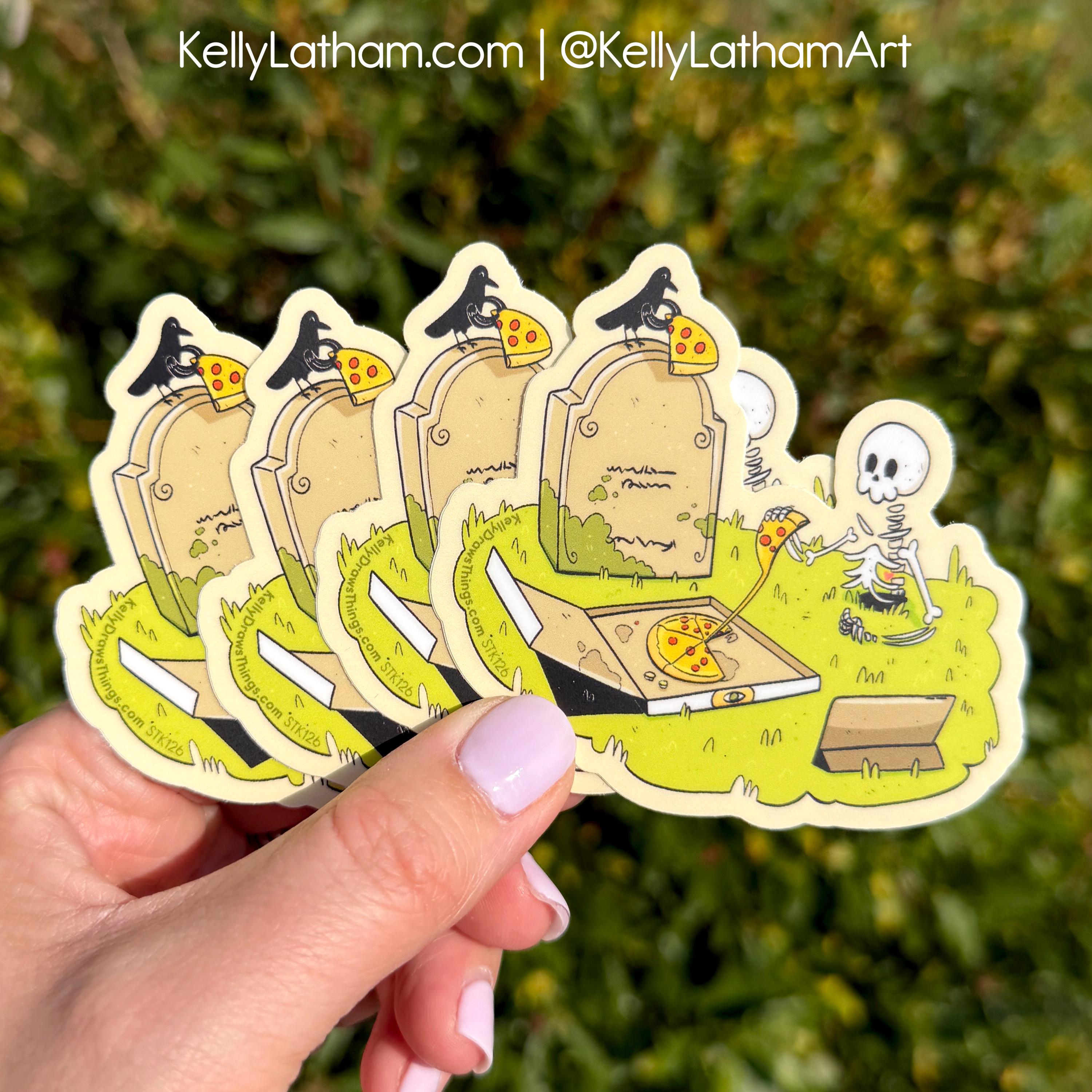 A hand holds a fanned-out set of die-cut stickers featuring a cartoon graveyard scene with tombstones, crows, a skeleton, and pizza slices, against a blurred green outdoor background. The text 'KellyLatham.