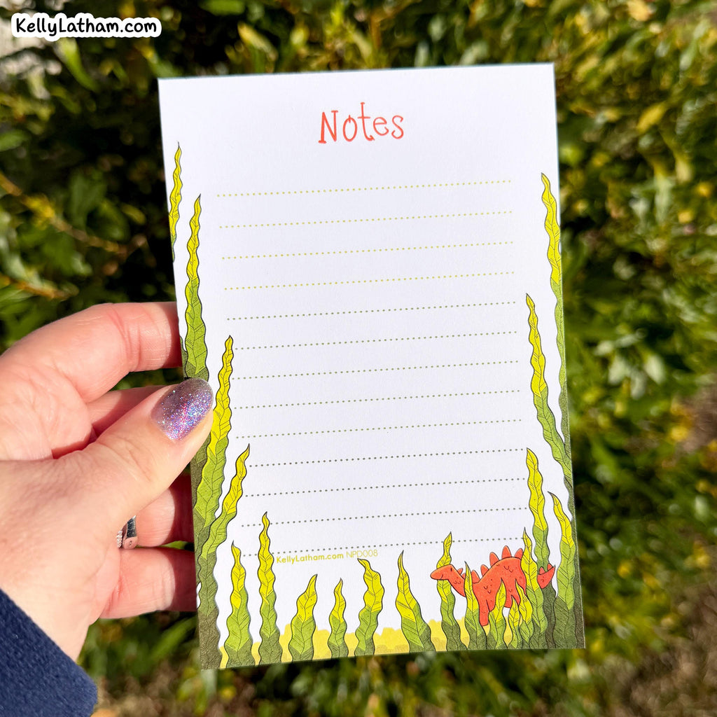 A hand holds a white notepad with a sea-plant illustration and the word 'Notes' at the top, photographed outdoors against blurred green foliage. The notepad has dotted writing lines and a small orange sea creature near the bottom.