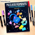 Mailbox Moments coloring book by Kelly Latham on a light wood desk, surrounded by colorful markers. The book cover features a black background with a blue backpack and colorful mailboxes floating out of it.