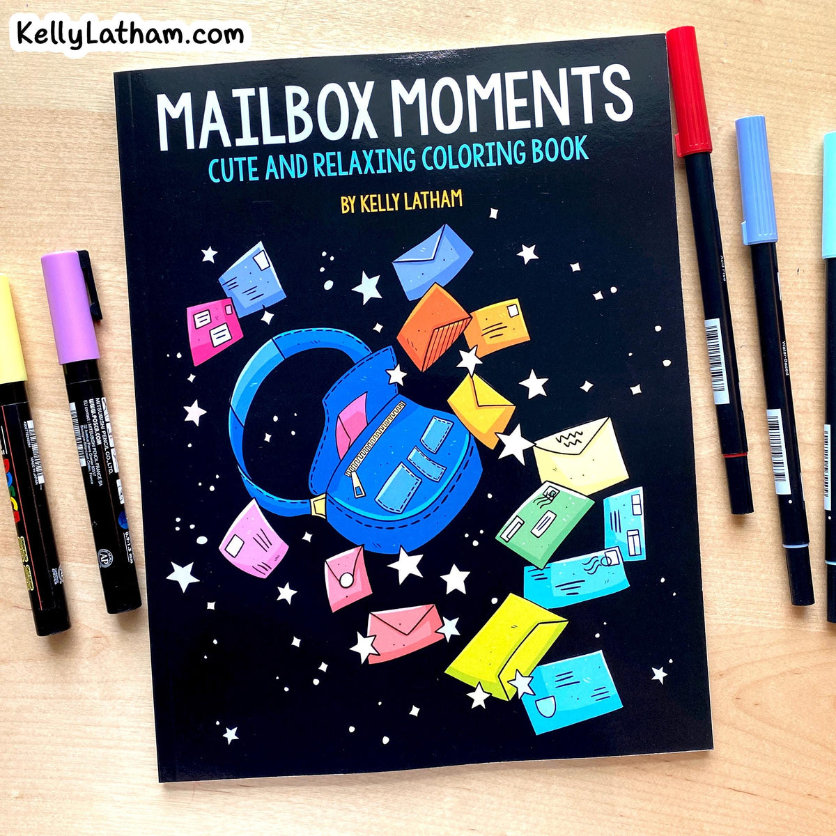 Mailbox Moments coloring book by Kelly Latham on a light wood desk, surrounded by colorful markers. The book cover features a black background with a blue backpack and colorful mailboxes floating out of it.