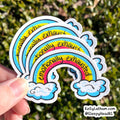 A hand holds a stack of five die-cut stickers featuring a rainbow with the text 'emotionally exhausted' in black script, set against a blurred green outdoor background.