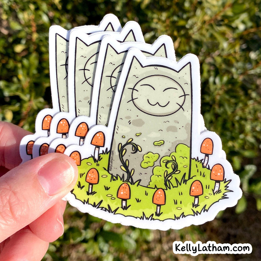 A hand holds a stack of die-cut stickers featuring a cute cartoon cat in a grassy field with orange mushrooms, photographed outdoors against blurred green foliage. A small 'KellyLatham.