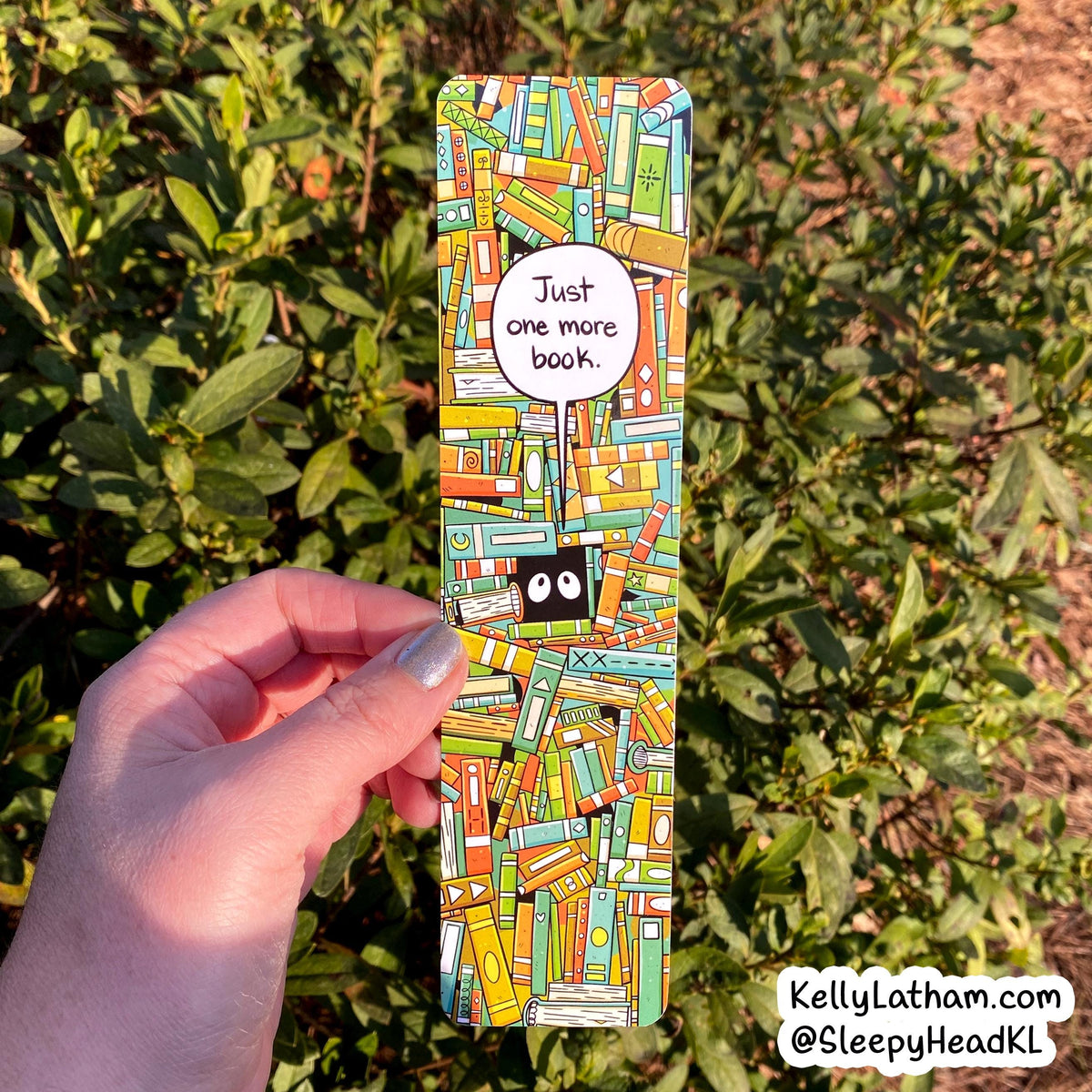 A hand holds a rectangular bookmark featuring a colorful pattern of illustrated books with a speech bubble that reads 'Just one more book.' The background is outdoor green foliage.