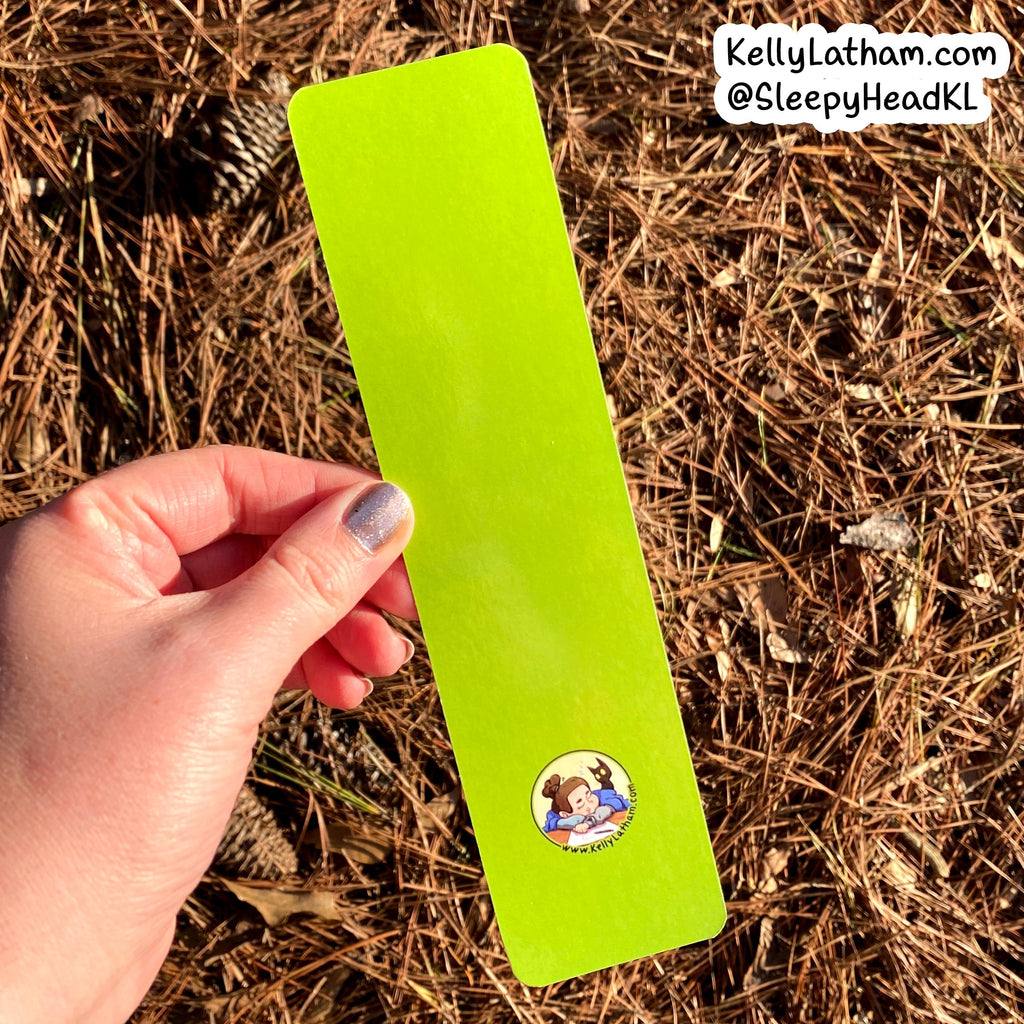 A hand holds a bright lime green rectangular bookmark or card outdoors over a bed of pine needles and pinecones. The card has a small round cartoon illustration near the bottom and a white text watermark in the top right corner.