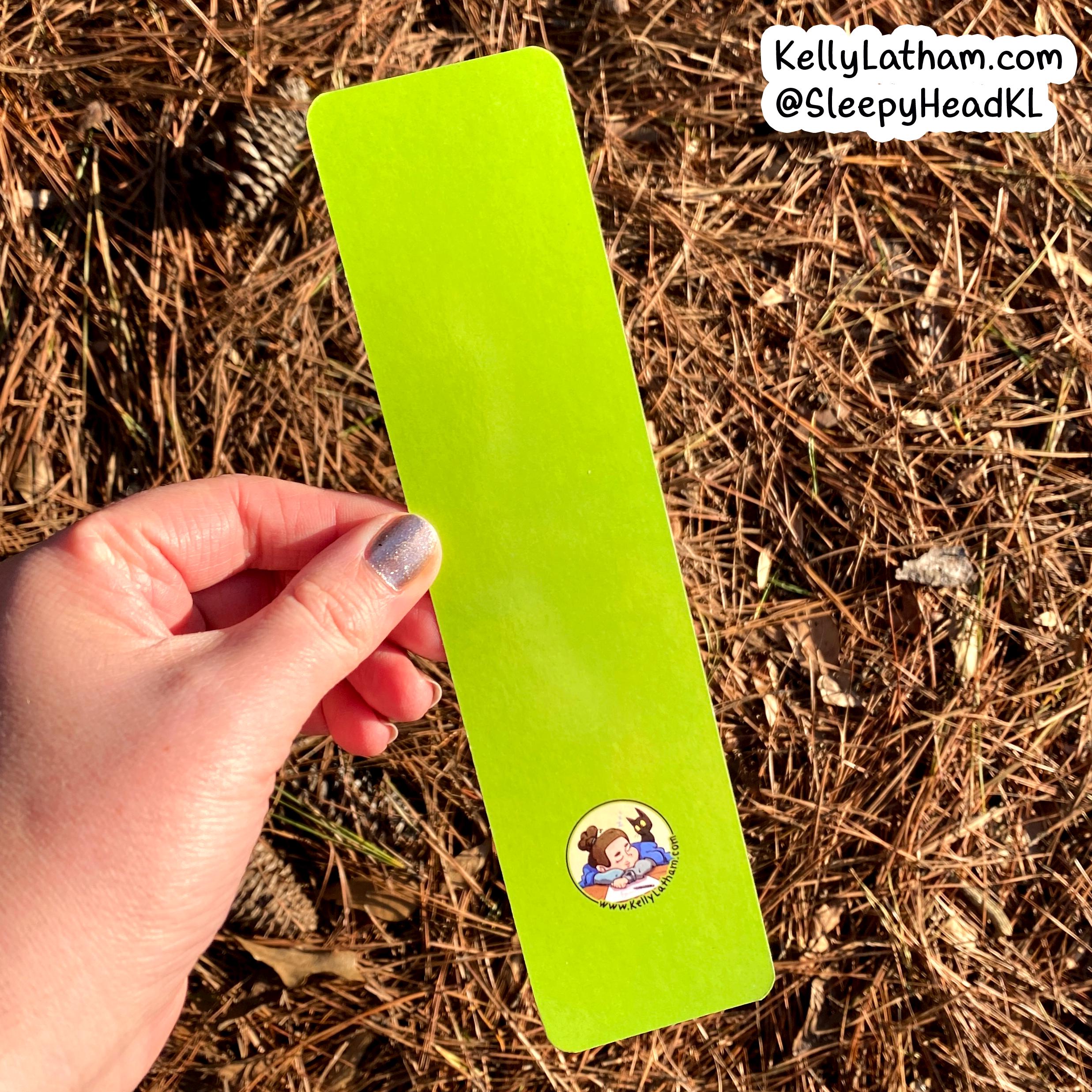 A hand holds a bright lime green rectangular bookmark or card outdoors over a bed of pine needles and pinecones. The card has a small round cartoon illustration near the bottom and a white text watermark in the top right corner.