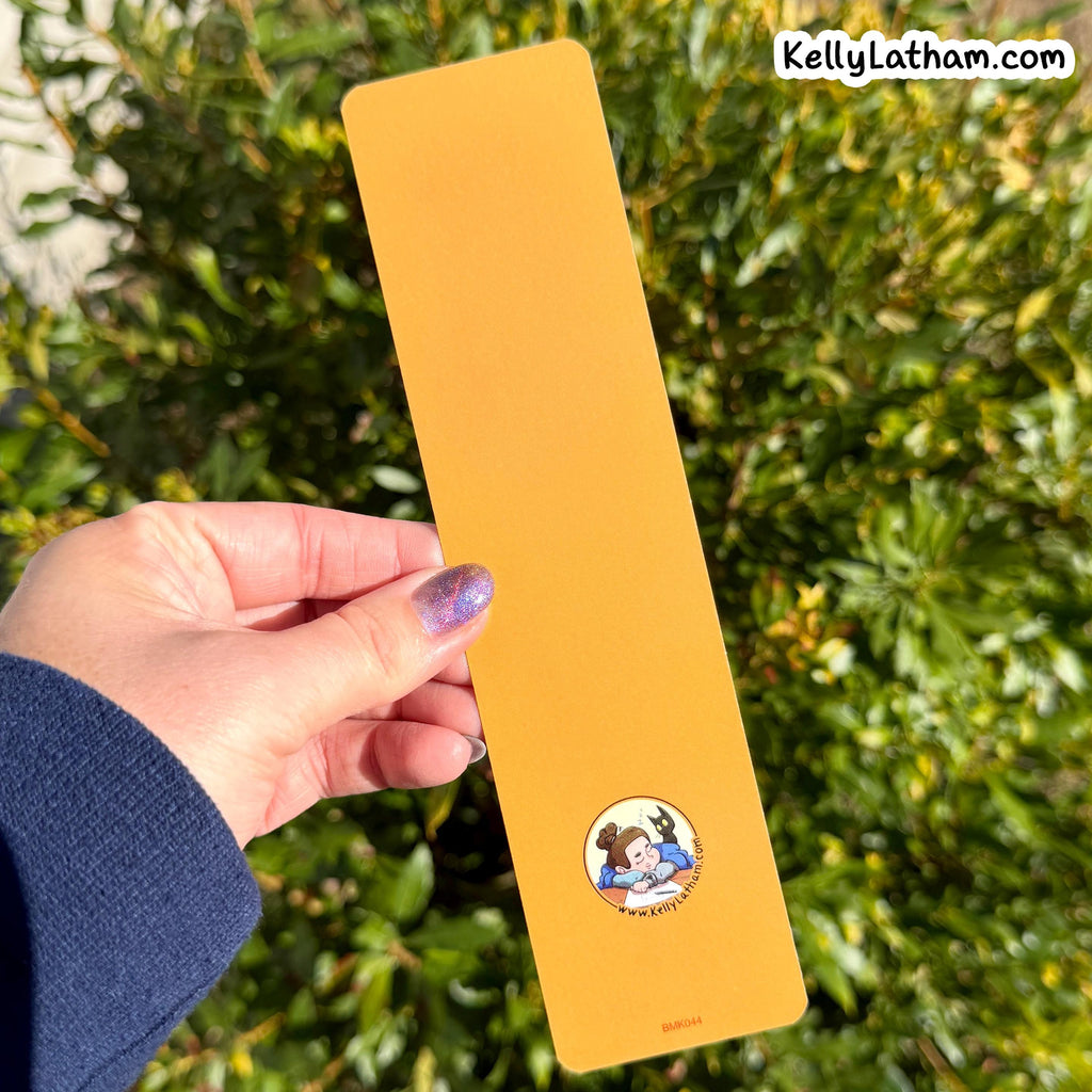A hand with purple glitter nail polish holds a long, rectangular mustard yellow card or bookmark outdoors in front of green foliage. The card features a small circular cartoon illustration near the bottom and the text 'KellyLatham.
