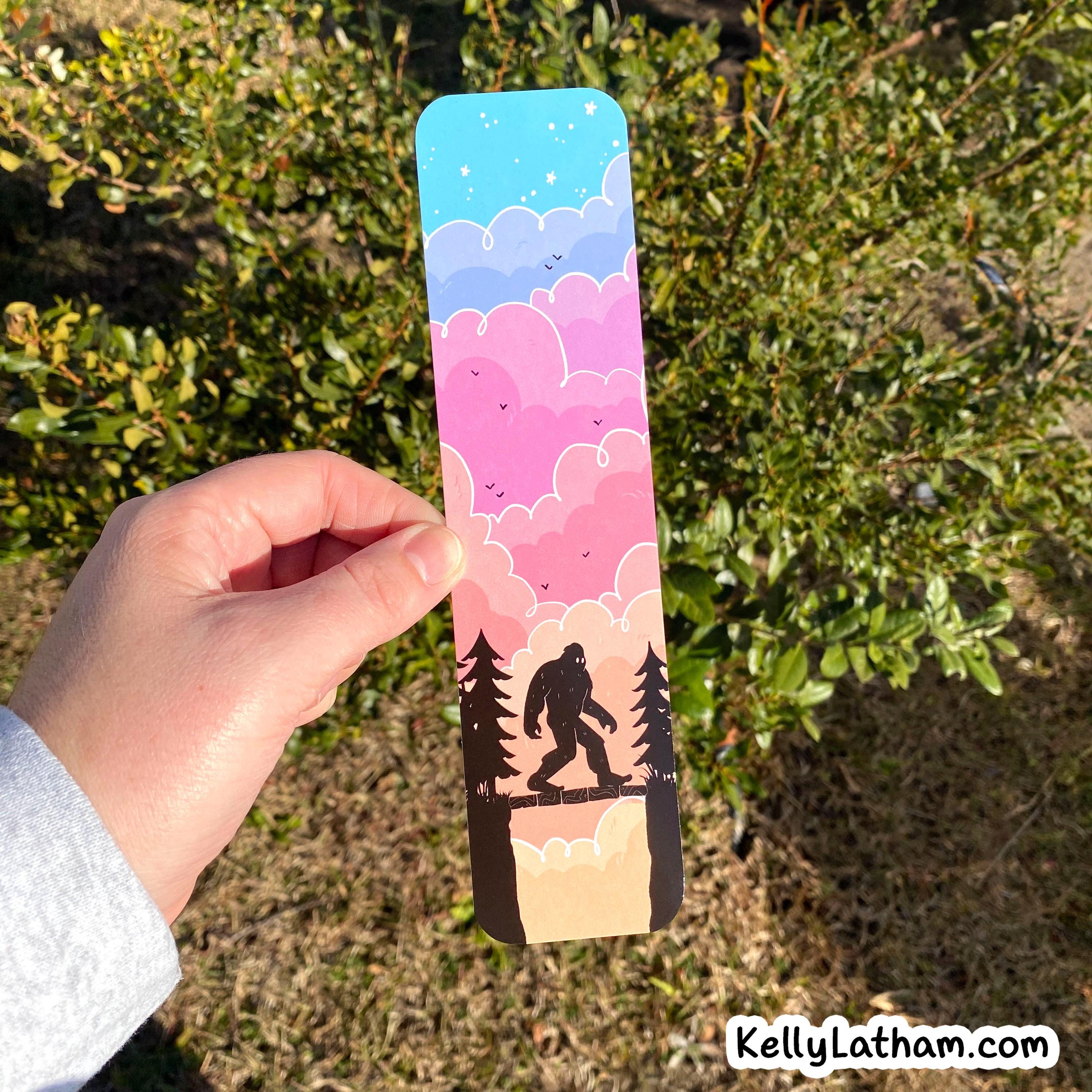 A hand holds a rectangular printed card or bookmark outdoors in front of green bushes. The printed design shows a night sky with blue, pink, and purple clouds, small birds, a silhouetted big-foot figure walking on a cliff, and dark pine trees.