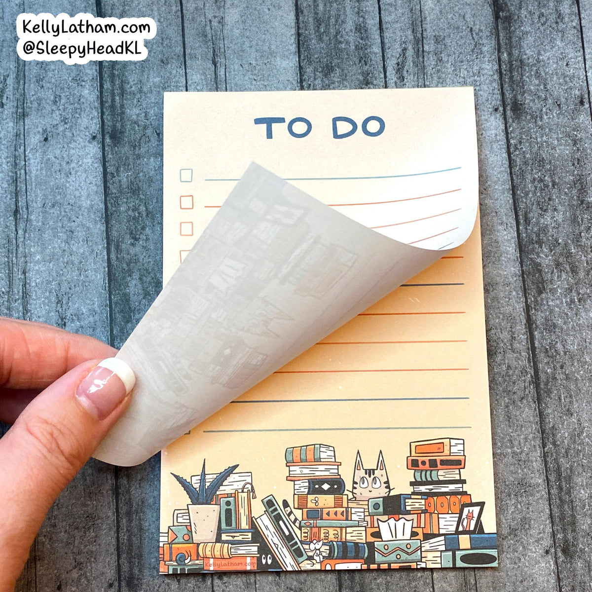 A hand with a French manicure holds a peeled-back translucent sticker sheet over a cream-colored notepad printed with the words 'TO DO' and a cute illustration of books, cassette tapes, and a cat at the bottom. The background is a gray wood plank surface, and a brand watermark is in the top-left corner.