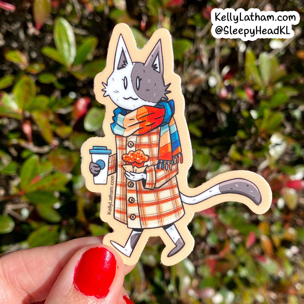 A die-cut sticker featuring an illustrated cat in a plaid coat and scarf, held by a hand with red nail polish against a blurred green and red outdoor background. The sticker has a visible brand watermark 'KellyLatham.
