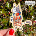 A die-cut sticker featuring an illustrated cat in a plaid coat and scarf, held by a hand with red nail polish against a blurred green and red outdoor background. The sticker has a visible brand watermark 'KellyLatham.