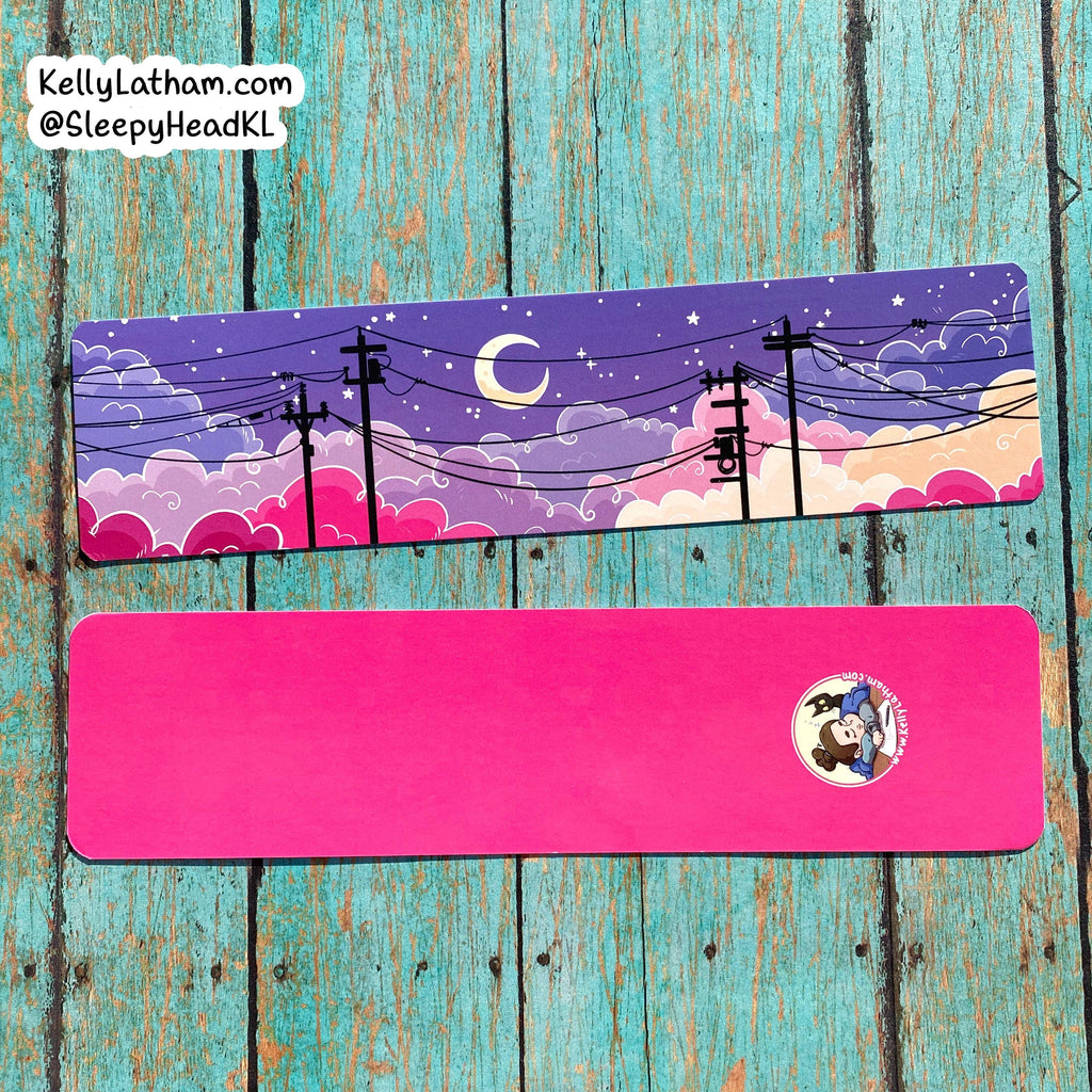 Two rectangular printed items laid flat on a distressed turquoise wood background. The top item shows a colorful night sky illustration with purple clouds, pink clouds, stars, a crescent moon, and black utility poles; the bottom item is bright pink with a small circular character print in the lower right.