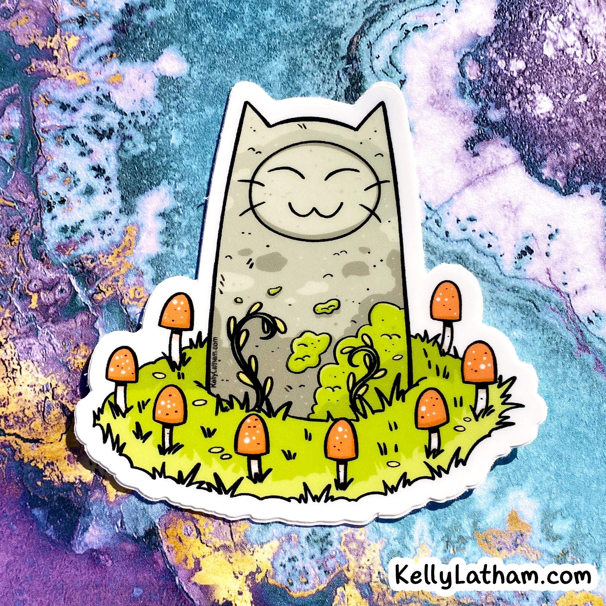 A die-cut sticker featuring a whimsical illustration of a gray cat sitting on a patch of green grass surrounded by orange mushrooms, shown against a colorful marbled background. The text 'KellyLatham.