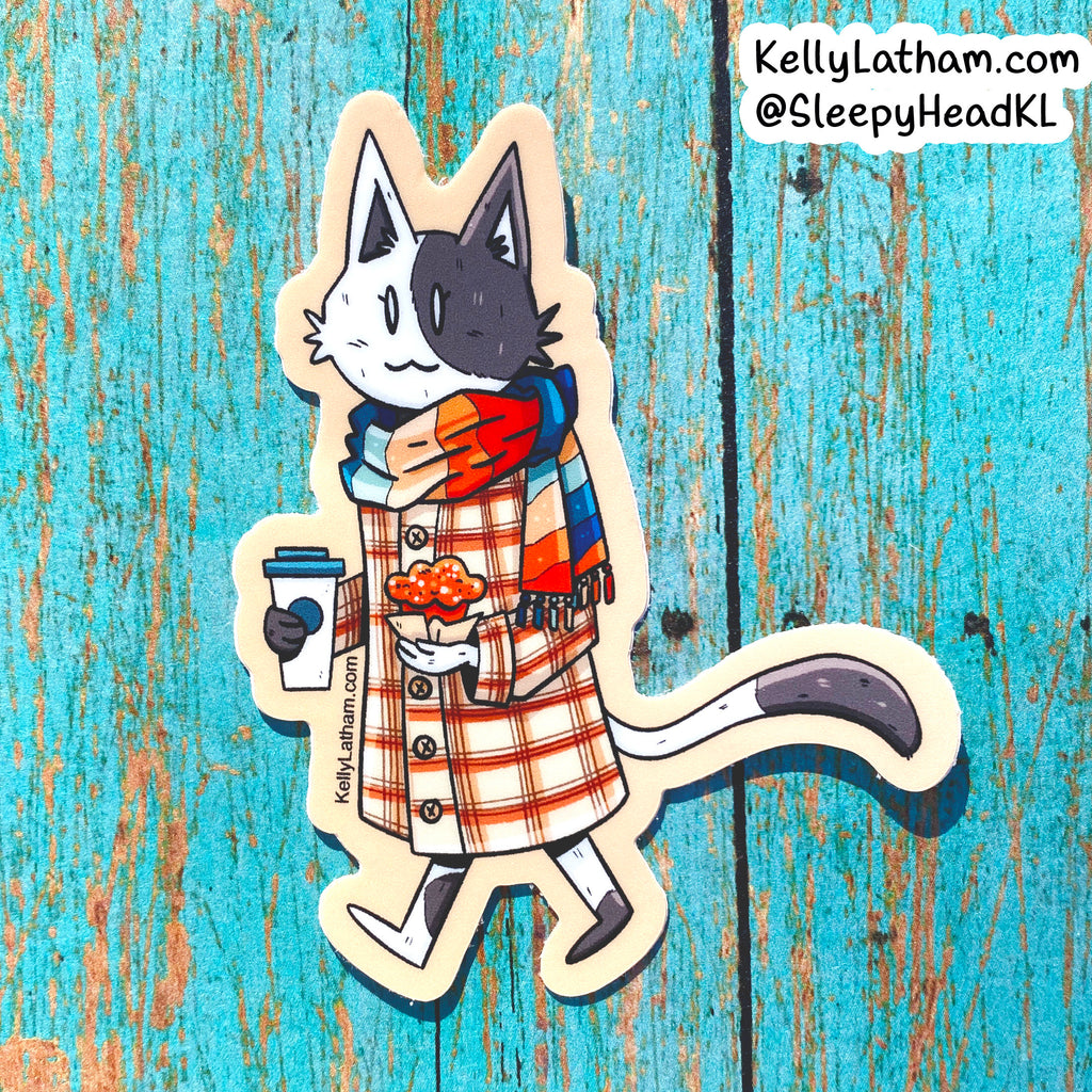 A die-cut sticker featuring an illustrated cat wearing a red and orange plaid coat and a blue, red, and orange striped scarf, holding a coffee cup and a snack, placed on a distressed turquoise wooden background. A brand watermark in the top right reads KellyLatham.