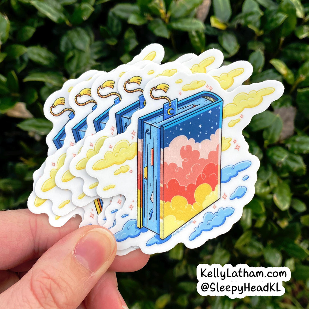 A hand holds a fanned stack of die-cut stickers featuring a colorful illustrated book with clouds and stars, set against a background of green foliage. A watermark in the bottom right corner reads KellyLatham.