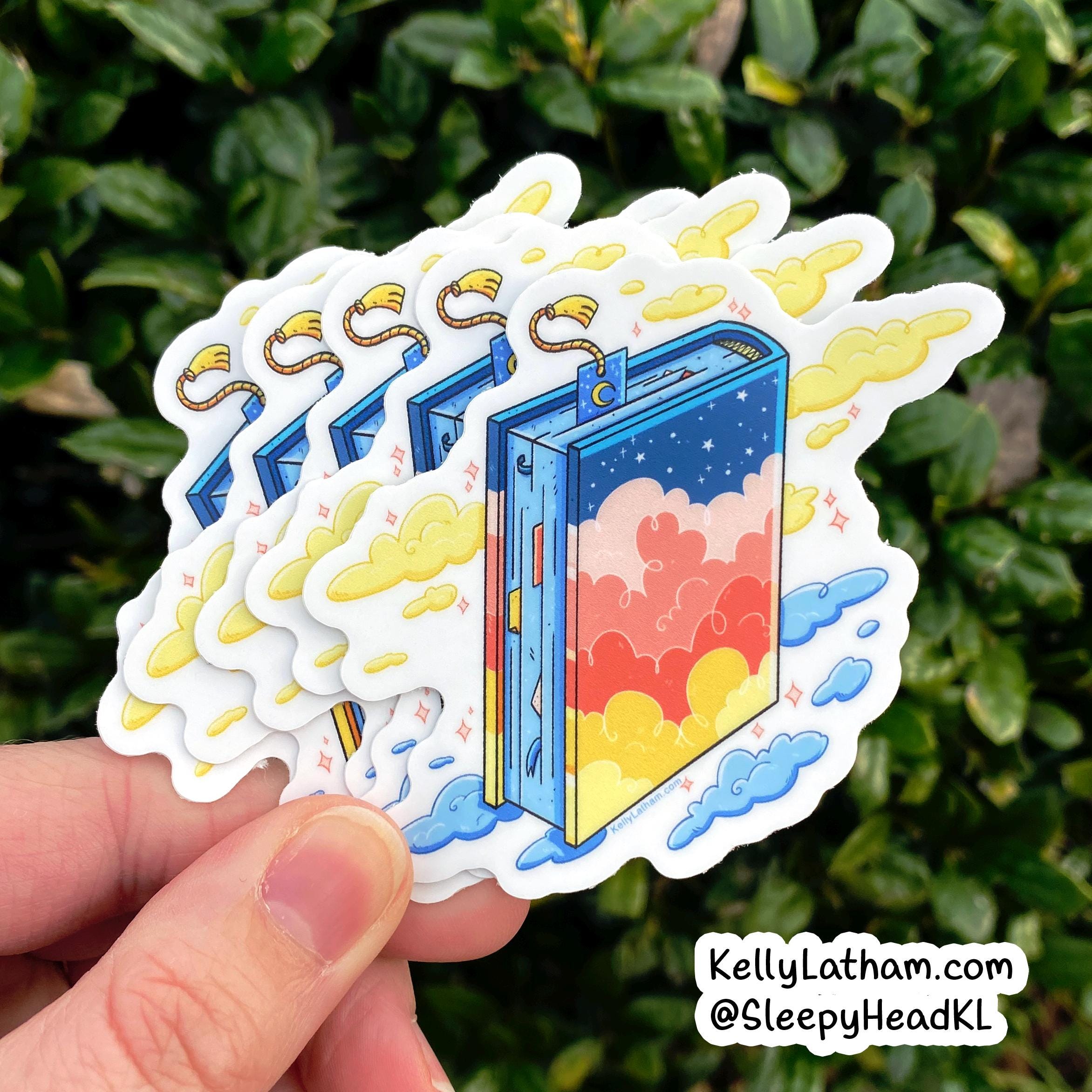 A hand holds a fanned stack of die-cut stickers featuring a colorful illustrated book with clouds and stars, set against a background of green foliage. A watermark in the bottom right corner reads KellyLatham.