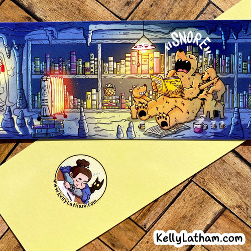 A printed illustrated strip or card showing a cartoon bear reading in a snowy library, placed on top of a pale yellow sheet with a circular cartoon logo and the text KellyLatham.com.