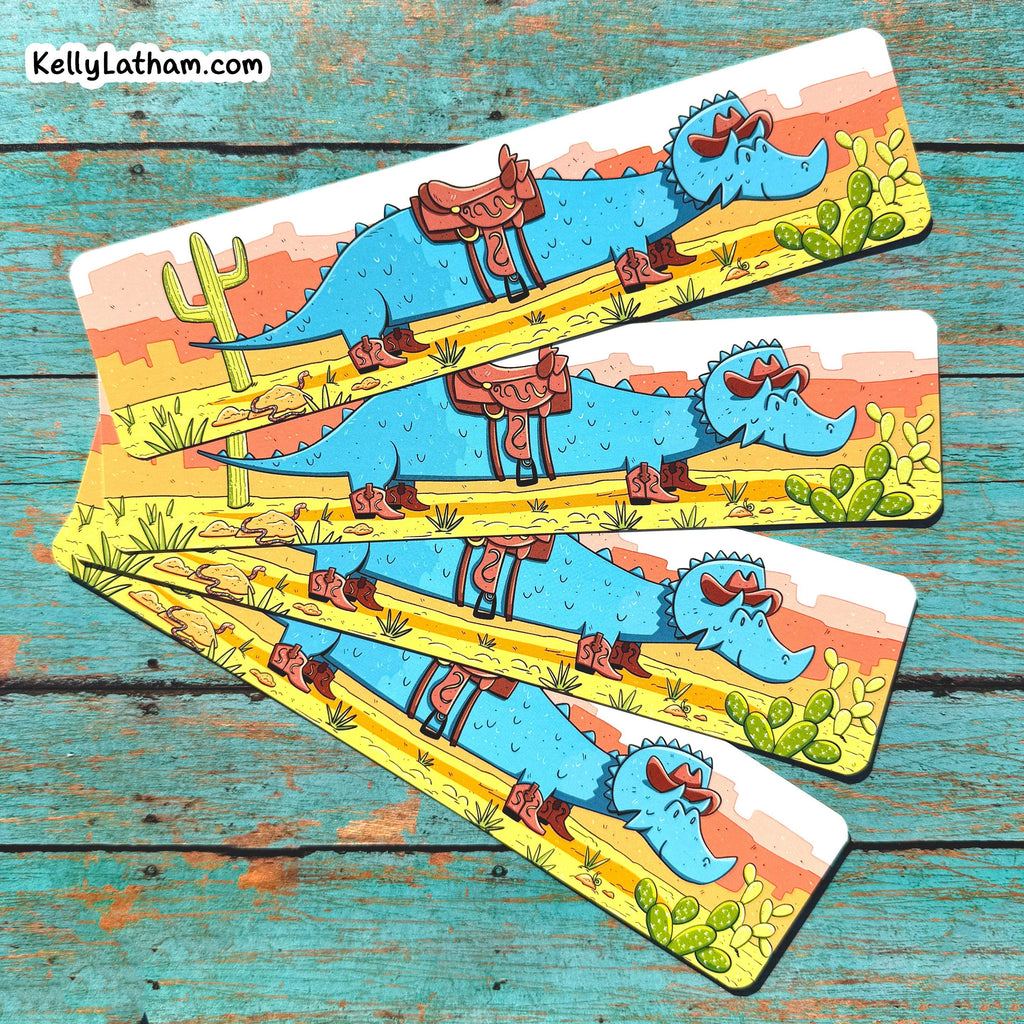 Four rectangular printed bookmarks fanned out on a distressed turquoise wooden surface. Each card shows a cartoon blue alligator wearing sunglasses and a brown saddle, with desert scenery including cacti and a yellow road.
