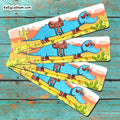 Four rectangular printed bookmarks fanned out on a distressed turquoise wooden surface. Each card shows a cartoon blue alligator wearing sunglasses and a brown saddle, with desert scenery including cacti and a yellow road.