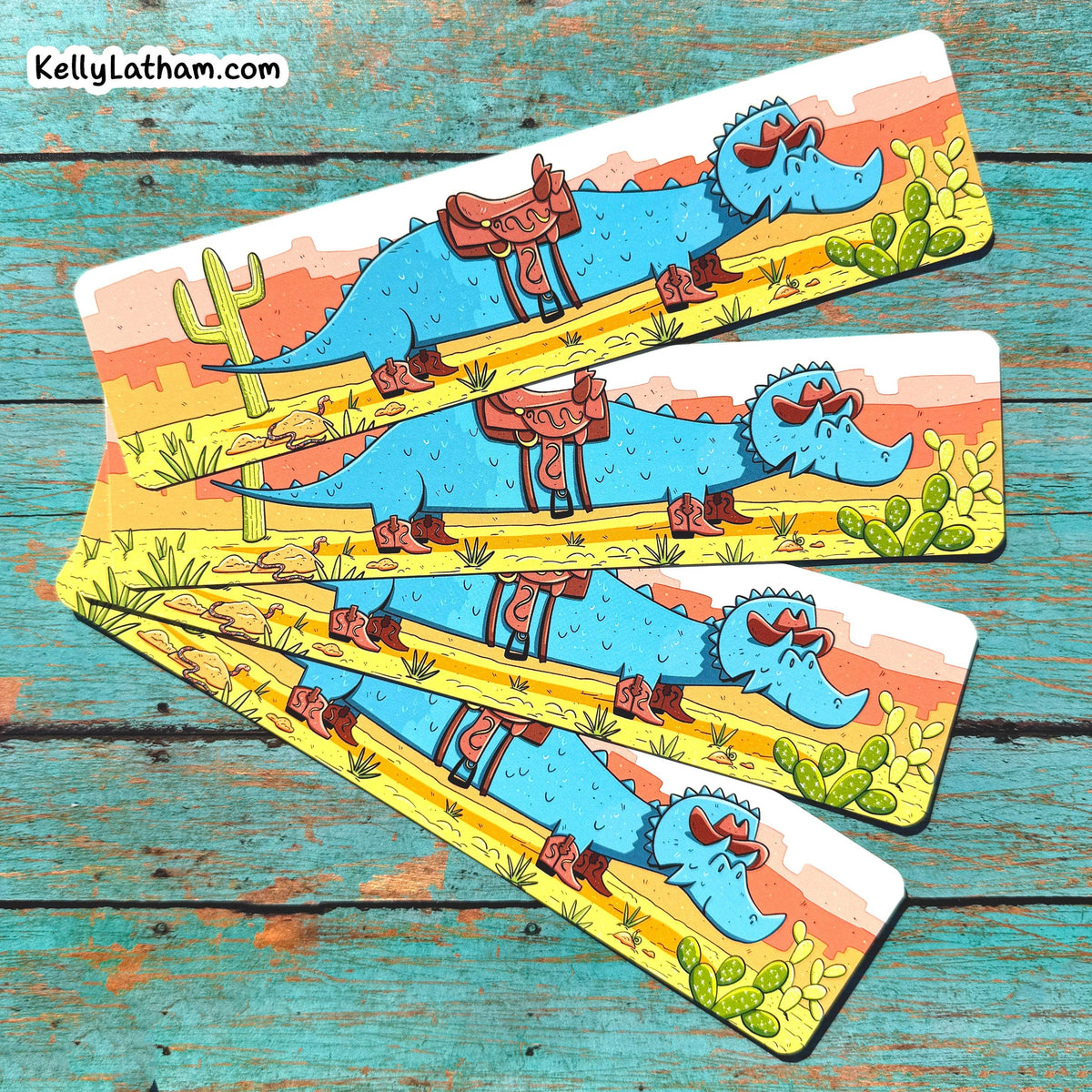 Four rectangular printed bookmarks fanned out on a distressed turquoise wooden surface. Each card shows a cartoon blue alligator wearing sunglasses and a brown saddle, with desert scenery including cacti and a yellow road.