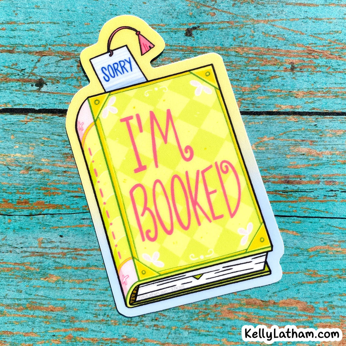 A die-cut sticker shaped like a yellow book with the words “I’M BOOKED” in pink script and a small tag reading “SORRY,” placed on a distressed turquoise wooden surface.