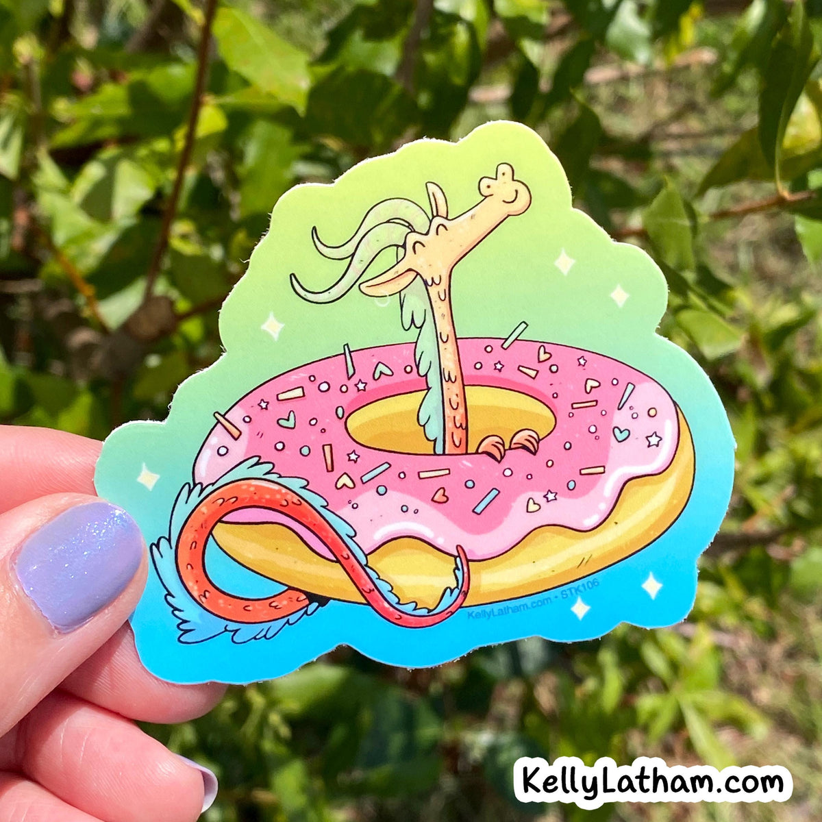 A hand with lavender nail polish holds a die-cut sticker featuring a whimsical illustration of a long-necked dragon rising from a pink frosted donut with sprinkles, set against a blurred green outdoor background.