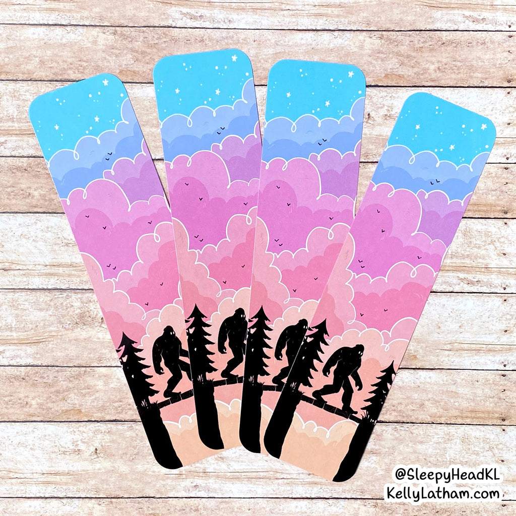 Four matching bookmarks with a whimsical pastel landscape design, displayed on a light wood background. The bookmarks feature a blue sky, pink and purple clouds, and black silhouettes of trees and Bigfoot figures walking along a river.
