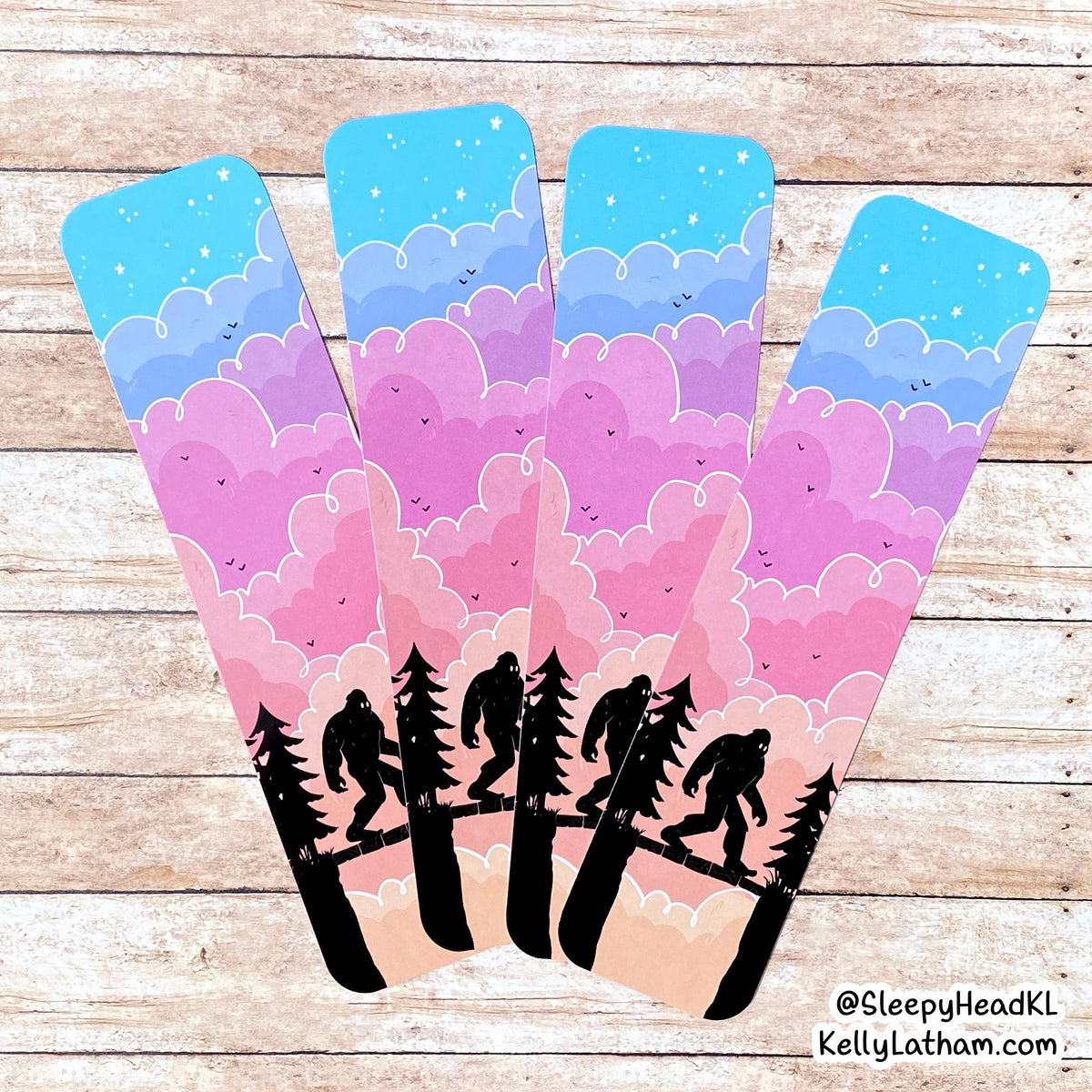 Four matching bookmarks with a whimsical pastel landscape design, displayed on a light wood background. The bookmarks feature a blue sky, pink and purple clouds, and black silhouettes of trees and Bigfoot figures walking along a river.