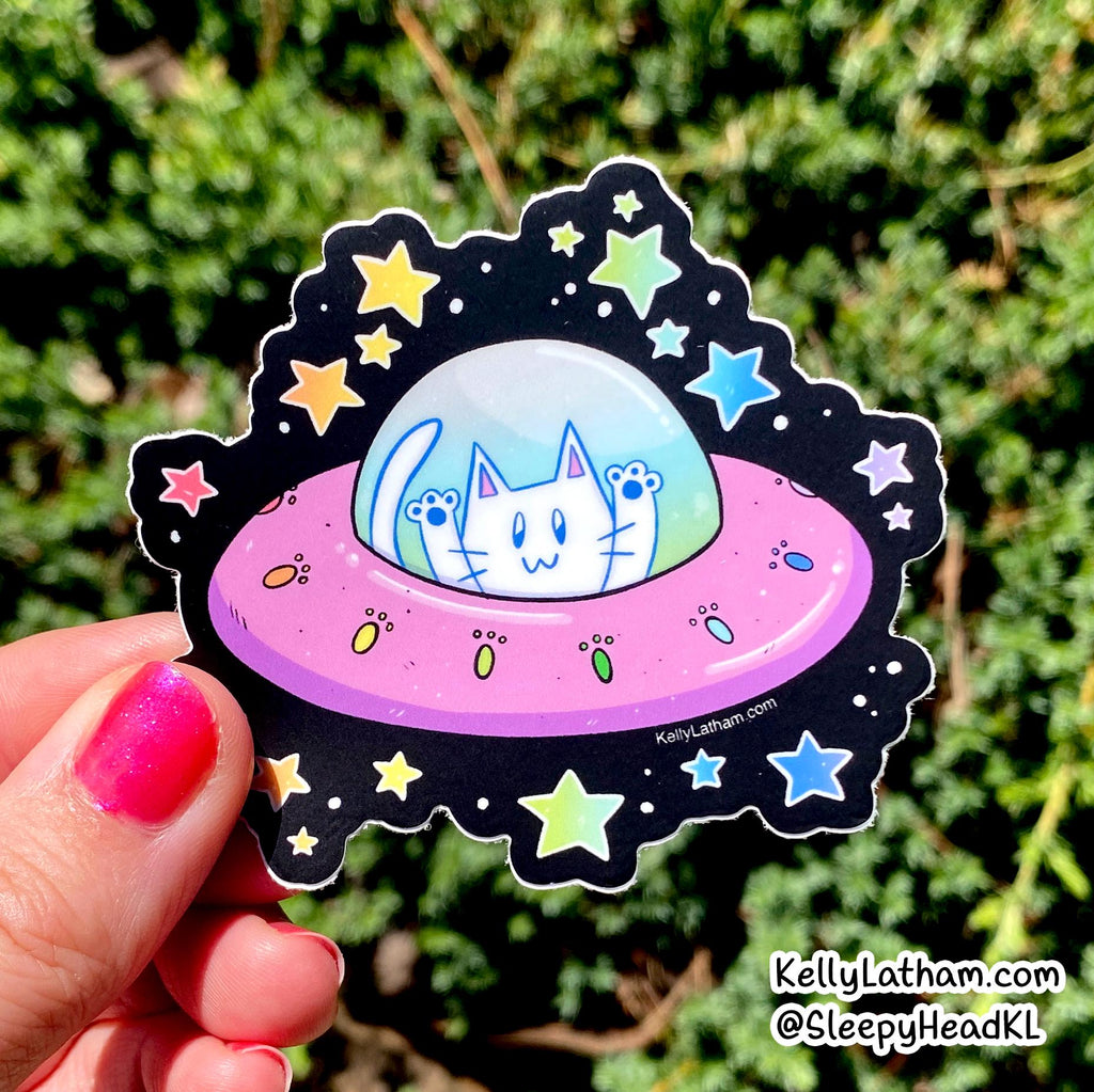 A hand with pink glitter nail polish holds a die-cut sticker featuring a white cat in a pink UFO surrounded by colorful stars on a black space background, with green foliage behind it.