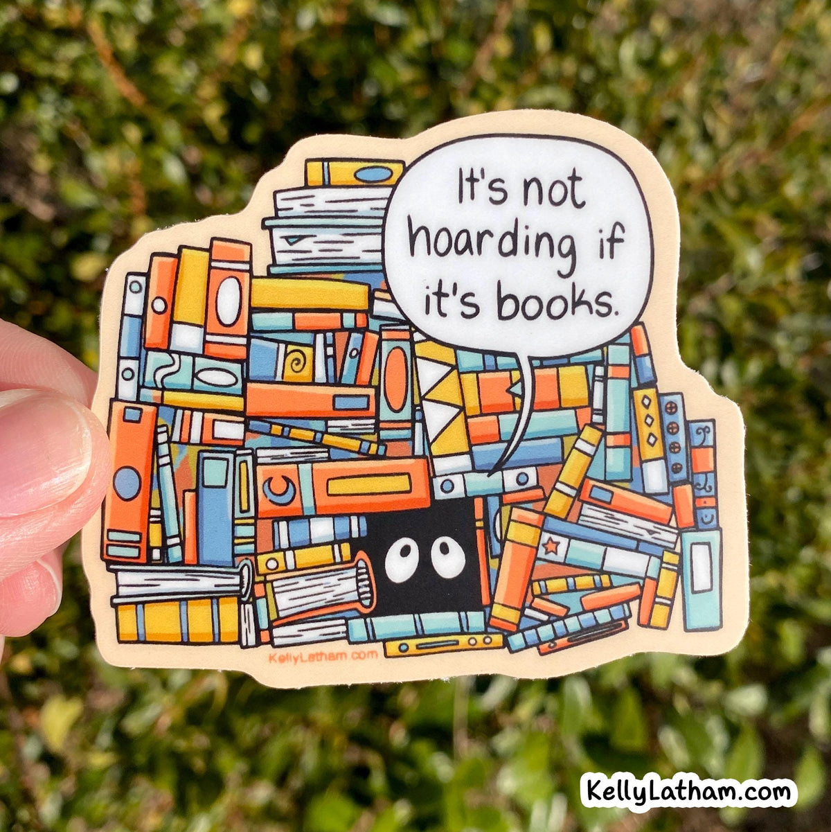 A hand holds a die-cut sticker featuring a colorful illustration of stacked books with a speech bubble that reads, 'It's not hoarding if it's books.' The background is blurred green foliage.