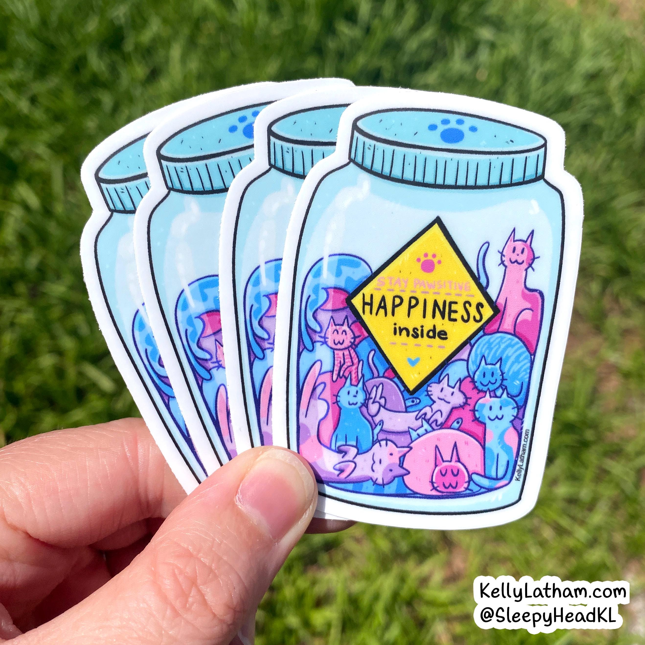 A hand holds four die-cut stickers shaped like light blue glass jars, fanned out against a blurred green grass background. The front sticker has a yellow diamond sign reading “HAPPINESS inside,” with colorful cartoon cats and small animals inside.
