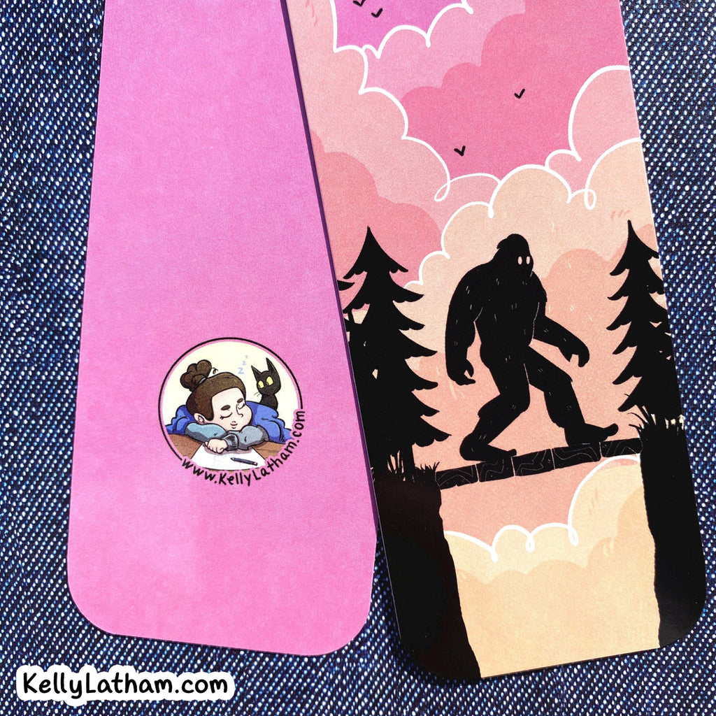 Two custom printed bookmarks or cards shown on a blue denim background. One card is solid pink with a circular cartoon illustration of bigfoot silhouette at sunset and the URL KellyLatham.