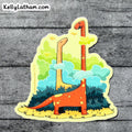 A die-cut sticker featuring a whimsical illustration of two long-necked dinosaurs standing among green bushes under a pale yellow sky, placed on a gray wood plank background. The sticker has the text 'KellyLatham.