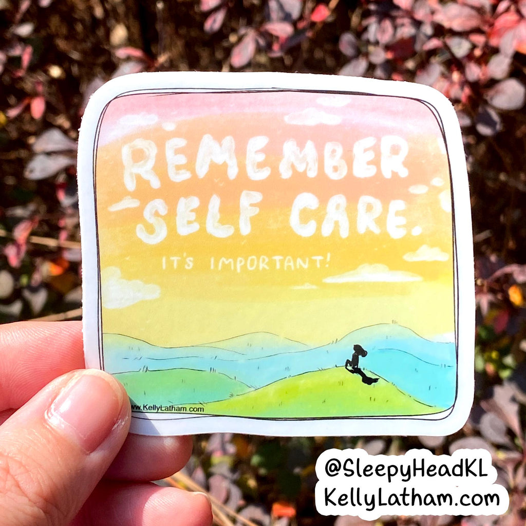 A hand holds a square sticker with rounded corners featuring a pastel landscape and the text 'REMEMBER SELF CARE IT'S IMPORTANT!'. The background shows blurred outdoor foliage.
