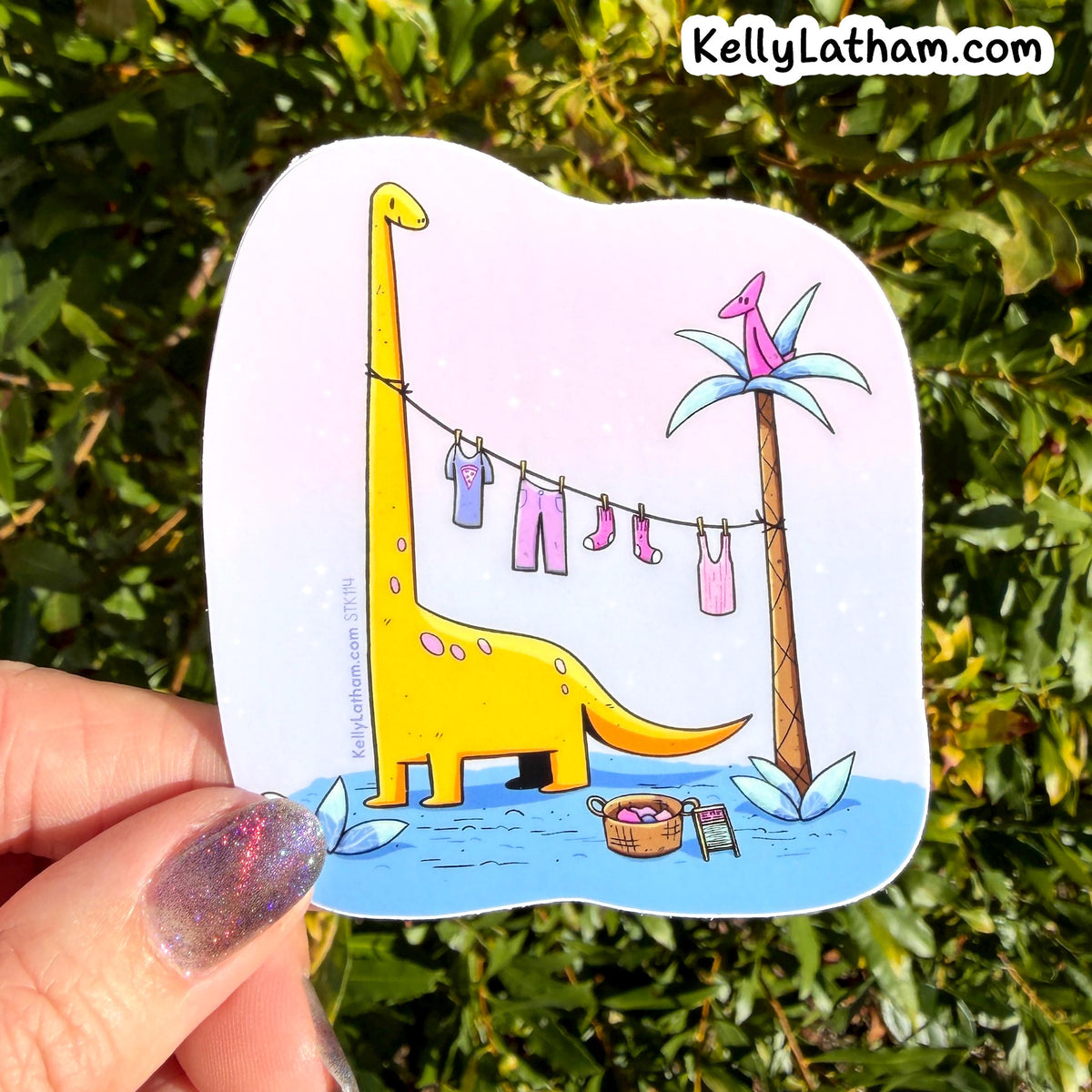 A hand with glittery nail polish holds a die-cut sticker featuring a yellow dinosaur laundry-drying scene, with a real green leafy background behind it. The sticker has the KellyLatham.