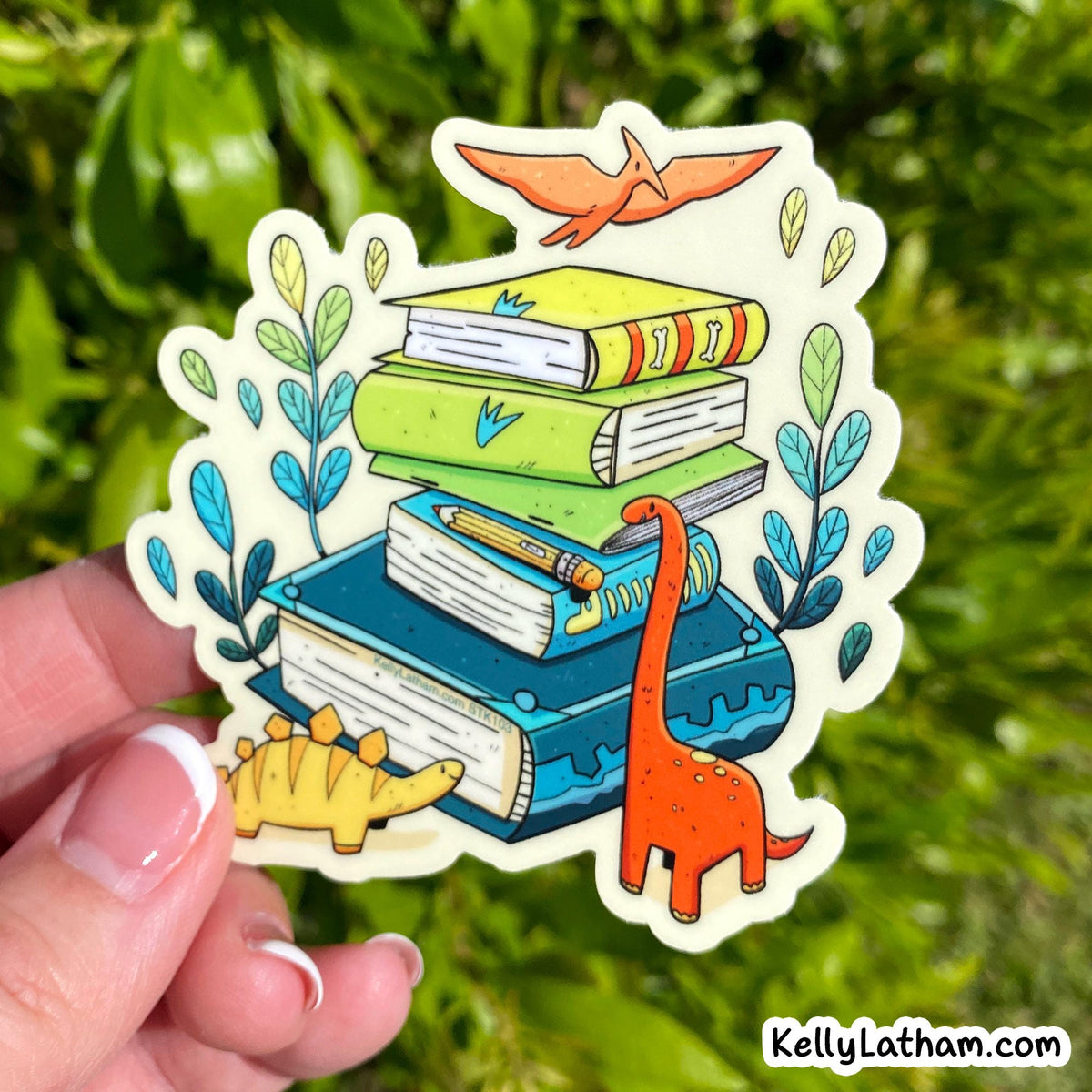A hand holds a die-cut sticker featuring a stack of colorful books with a pterodactyl, ferns, and two dinosaurs, set against a blurred green outdoor background.